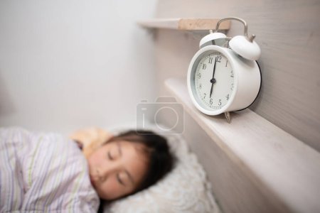 Foto de Pequeña chica asiática durmiendo. despertador en el estante - Imagen libre de derechos
