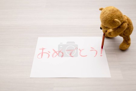 Photo for Teddy bear writing congratulations in Japanese with crayons on paper - Royalty Free Image