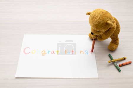 Foto de Osito de peluche escribiendo 'felicitaciones' en papel con lápices de colores - Imagen libre de derechos