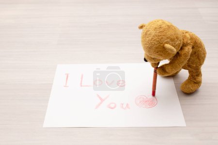 Téléchargez les photos : Ours en peluche écrivant "Je t'aime" sur papier avec un crayon - en image libre de droit