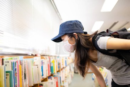 Foto de Mujer con una máscara eligiendo un libro en la biblioteca - Imagen libre de derechos