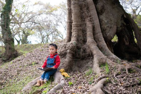 Téléchargez les photos : Jeune asiatique enfant assis dans un grand arbre avec livre - en image libre de droit