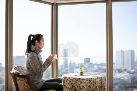 Foto de Una mujer tomando té junto a la ventana de un apartamento de gran altura - Imagen libre de derechos