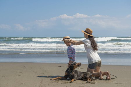Photo for Parent and child and a Doberman relax on the beach - Royalty Free Image