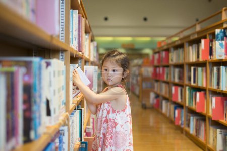 Téléchargez les photos : Asiatique petite fille choisir livre à la bibliothèque - en image libre de droit