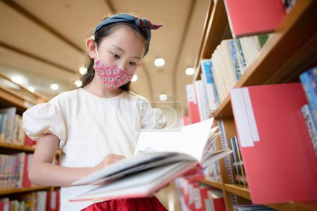 Photo for Little girl in mask reading book in library - Royalty Free Image