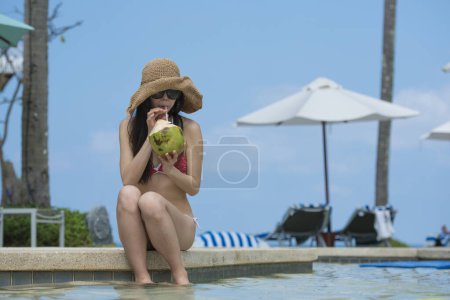 Téléchargez les photos : Belle japonaise femme boire du jus de noix de coco sur le bord de la piscine - en image libre de droit
