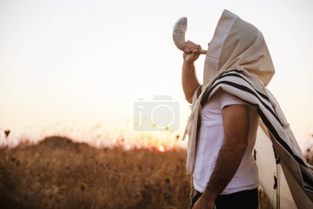 Photo for Jewish man in a traditional tallit prayer shawl blowing the ram's horn shofar, in the field against sunrise sky on Elul, Rosh HaShana and Yom Kippurim - Royalty Free Image