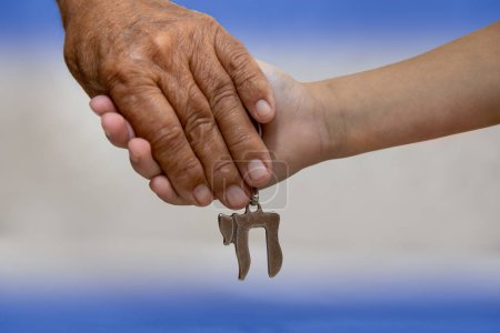 Photo for Am Israel Chai. Hands of an elderly man holding the hand of a child close up. they holding a silver Chai key chain, which spell "life" in hebrew. blue and white (Israel flag) background. - Royalty Free Image