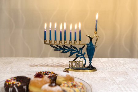 Photo for Beautiful Menorah (Chanukkiah) with 8 lit burning candles for Jewish Hanukkah holiday on table at home. Celebrating Chanukah festival of lights. Sufganiyot donuts sweet cultural food on a plate. - Royalty Free Image