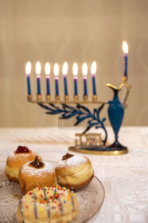 Photo for Beautiful Menorah ,Chanukkiah with 8 lit burning candles for Jewish Hanukkah holiday on table. Celebrating Chanukah festival of lights. Sufganiyot donuts sweet cultural food on a plate in focus - Royalty Free Image