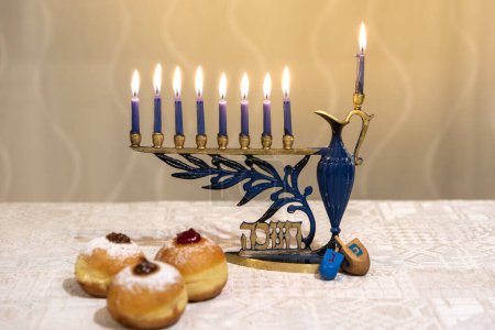 Photo for Menorah (Chanukkia) with 8 lit burning candles for Jewish Hanukkah holiday on table at home. Celebrating Chanukah festival of lights. Dreidel and Sufganiyot donuts sweet cultural food on a plate. - Royalty Free Image