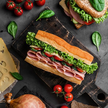 Photo for Delicious baguette sandwich with ham, bacon, cheese, lettuce, tomatoes, sausage, gammon on cutting board with herb and spices over on dark background. Meat food, top view, flat lay, toning - Royalty Free Image
