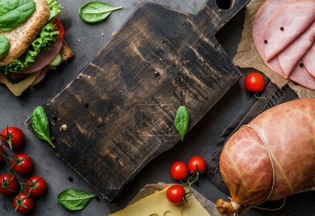 Photo for Wooden cutting board on dark background witn delicious sandwich, ham, bacon, cheese, spices, tomatoes, sausage, gammon, herb. Meat food, top view, toning, place for text - Royalty Free Image