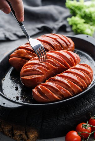 Téléchargez les photos : Saucisses fumées grillées dans une casserole en bois foncé avec tomates et épices. Saucisse chaude à la fourchette, aliments à base de viande, mise au point sélective - en image libre de droit