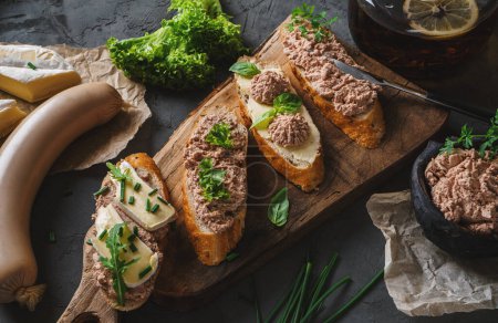 Foto de Deliciosos sándwiches con paté, queso, verduras, especias en la tabla de cortar con cebolla sobre fondo oscuro. Comida de carne, vista superior, tonificación - Imagen libre de derechos