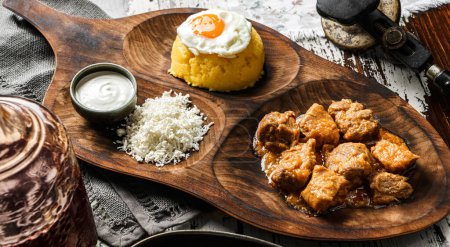 Photo for Delicious polenta with fried eggs, cheese, stew meat and sour cream on wooden board over rustic background. Traditional romanian healthy food, top view selective focus - Royalty Free Image