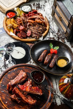 Photo for Set of meat dishes, fried sausages on a plate, grilled meat and pig ears with fries potatoes on cutting board, barbecue pork spare ribs with hot honey chilli sauce on wooden table. Meat food, top view - Royalty Free Image