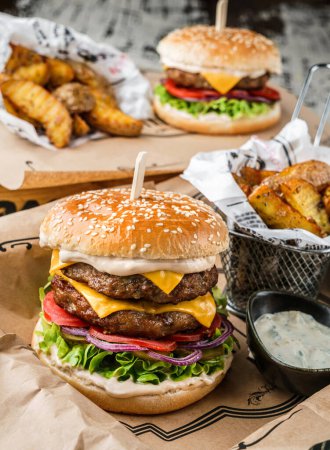 Téléchargez les photos : Burger de boeuf et double burger avec laitue, tomates, tranche de fromage, cornichons, sauce, rondelles d'oignons, frites sur papier artisanal sur fond bois, gros plan - en image libre de droit