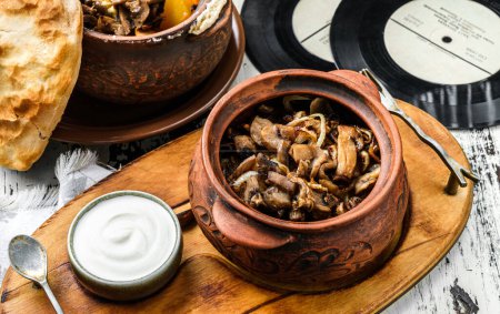 Foto de Las setas cocidas con la cebolla en la olla de barro con la crema agria a la tabla de madera. Comida casera, de cerca - Imagen libre de derechos