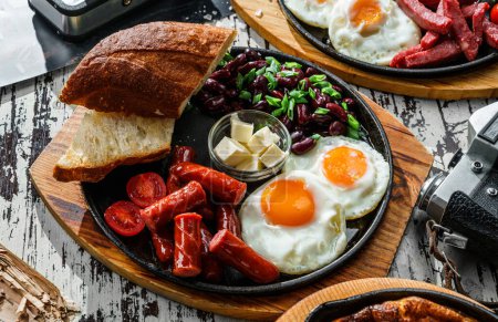 Photo for Delicious breakfast with fried eggs, slices of bread, sausage and salad of beans served on hot pan over wooden background. Healthy breakfast, close up - Royalty Free Image