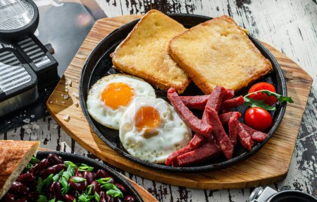 Photo for Delicious breakfast with fried eggs, toasts, sausage and vegetables served on hot pan over wooden background. Healthy breakfast, close up - Royalty Free Image