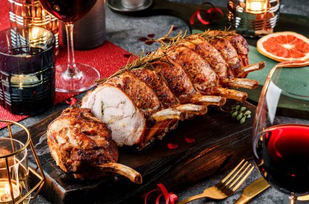 Photo for Holiday baked meat tenderloin with ribs, pork, roast beef on dark wooden board with glasses, bottle of wine, candles, festive decoration. Christmas and New Year food, bokeh, lights - Royalty Free Image