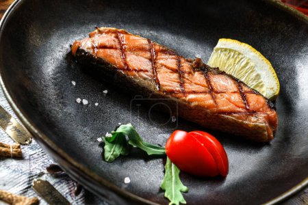 Foto de Filete de salmón a la parrilla con rodajas de limón y tomates en plato sobre fondo rústico de madera. Mariscos, comida sana, primer plano - Imagen libre de derechos