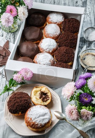 Photo for Yummy set of cake donuts with vanilla, chocolate icing, powdered sugar and chocolate chips in paper box on grey background with flowers. Sweets, dessert and pastry, top view - Royalty Free Image