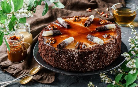 Photo for Chocolate cake with caramel, condensed milk, cream and walnuts on dark background with spring flowers. Sweets, dessert and pastry, top view - Royalty Free Image