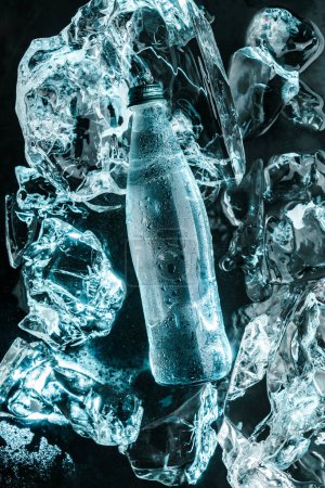 Téléchargez les photos : Bouteille d'eau dans la glace. Glace éclatée de lumière bleue. Publicité eau minérale en bouteille en verre, vue de dessus - en image libre de droit