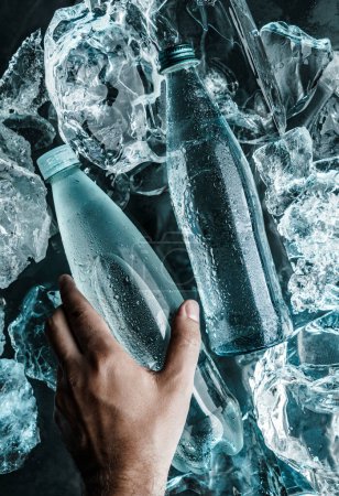 Téléchargez les photos : Un homme tenant une bouteille d'eau dans la glace. Glace éclatée de lumière bleue. Publicité eau minérale en bouteille en verre, vue de dessus - en image libre de droit
