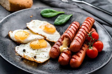 Photo for Delicious breakfast with fried eggs, grilled sausages with cheese, tomatoes, greens and spices on plate on dark background. Meat food, selective focus - Royalty Free Image