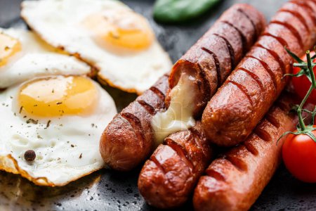 Photo for Delicious breakfast with fried eggs, grilled sausages with cheese, tomatoes, greens and spices on plate on dark background. Meat food, selective focus - Royalty Free Image