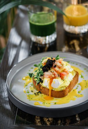 Photo for Delicious toast with egg Benedict or poached egg, black caviar, shrimps and microgreens on plate on marble table with smoothies. Healthy breakfast food, diet, top view - Royalty Free Image