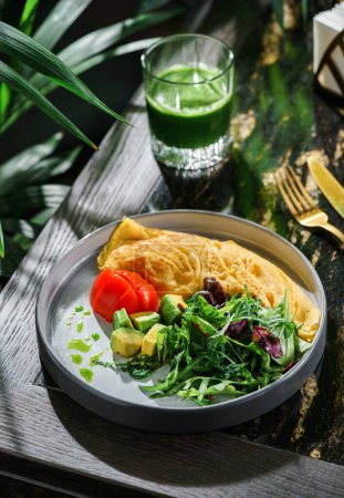 Photo for Omelette with fresh salad, arugula, avocado and tomato on plate on table with green smoothie. Healthy breakfast food, morning light, top view - Royalty Free Image