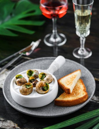 Foto de Deliciosos caracoles marinos con hierbas y pan en plato sobre fondo de mármol con bebidas. Comida gourmet. Escargot Caracoles, vista superior - Imagen libre de derechos