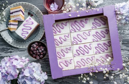 Téléchargez les photos : Des morceaux de gâteau mousse crème avec des baies en boîte et sur l'assiette sur fond gris clair avec des baies et des fleurs violettes. Bonbons, dessert et pâtisserie, vue sur le dessus - en image libre de droit
