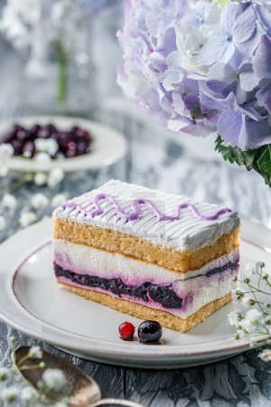 Téléchargez les photos : Pièce de gâteau mousse crème avec des baies sur fond gris clair avec des baies et des fleurs violettes. Bonbons, dessert et pâtisserie, vue sur le dessus - en image libre de droit