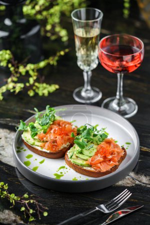 Photo for Sandwiches with cream cheese, avocado, slices salmon, red fish caviar and microgreens on plate over table with drinks. Healthy breakfast food, top view - Royalty Free Image