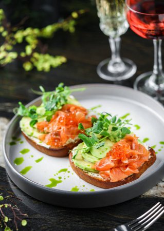 Photo for Sandwiches with cream cheese, avocado, slices salmon, red fish caviar and microgreens on plate over table with drinks. Healthy breakfast food, top view - Royalty Free Image