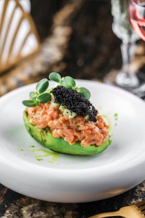 Foto de Delicioso tártaro de aguacate y salmón crudo con caviar negro en plato sobre mesa de mármol con copas de vino. Comida saludable, primer plano, vista superior - Imagen libre de derechos