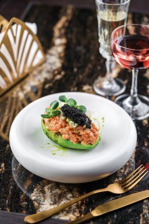 Photo for Delicious avocado and raw salmon fish tartare with black caviar on plate on marble table with wine glasses. Healthy food, close up, top view - Royalty Free Image