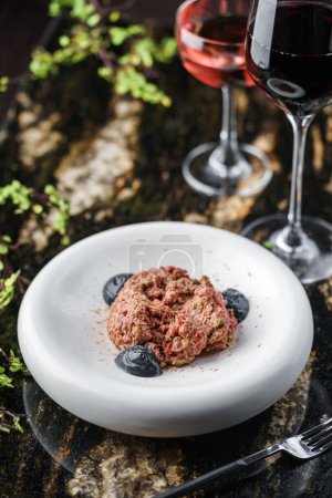 Photo for Delicious beef meat tartare with pickled cucumber and truffle sauce on plate on marble table with wine glasses. Healthy food, close up, top view - Royalty Free Image