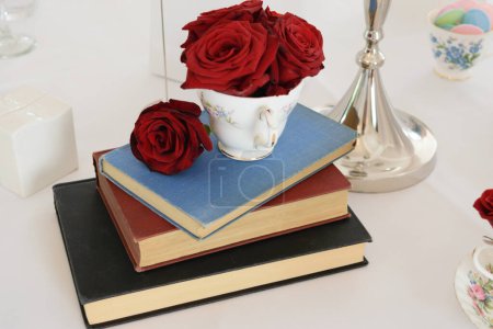 A stack of three books with a teacup filled with red roses on top, placed on a white tablecloth.