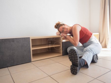 A cheerful young woman rests next to her newly assembled wooden furniture, showcasing a sense of achievement and relaxation