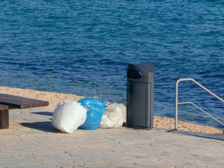 Basura desbordante junto a un cubo lleno en un paseo marítimo, destacando la necesidad de una gestión regular de los residuos en las zonas costeras
