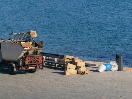 Ein Müllabfuhrwagen, der auf einer Strandpromenade geparkt ist, umgeben von Haufen von Pappe, Tüten und anderem Müll, der auf Umweltbedenken hinweist