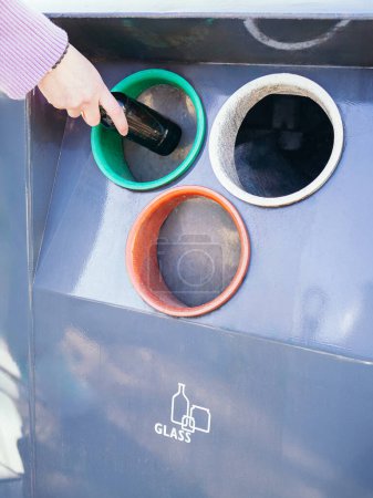 Nahaufnahme einer Hand, die eine dunkle Glasflasche in einen Recyclingbehälter mit Farbcodierung legt, der für Glas bestimmt ist, um die ökologische Verantwortung zu fördern