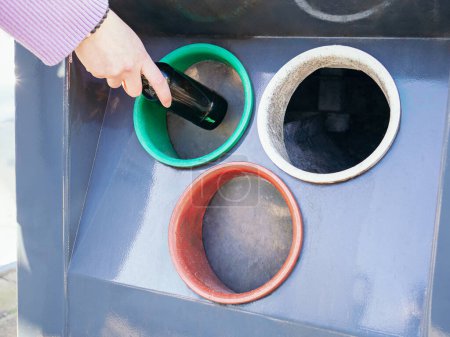 Die Hand einer Person wird dabei gesehen, wie sie eine dunkle Glasflasche in einen bunten Recyclingbehälter entsorgt, was auf umweltfreundliche Praktiken hinweist